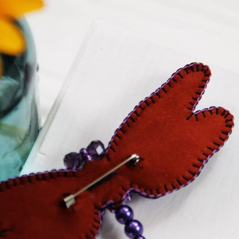 May include: The back of a purple and red beaded dragonfly brooch with a silver pin. The dragonfly has a red felt body and purple beaded wings.