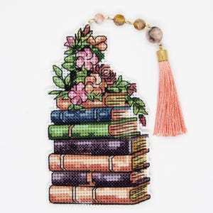Könnte beinhalten: Ein Kreuzstich-Lesezeichen mit einem Bücherstapel, der mit Blumen verziert ist. Das Lesezeichen hat eine rosa Quaste und eine Perlenkette. Die Bücher sind in Blau-, Grün-, Lila- und Orangetönen gehalten.