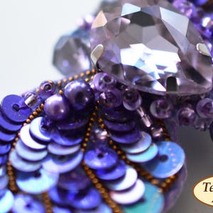 May include: A purple and blue beaded dragonfly brooch with a large iridescent crystal in the center. The dragonfly's wings are made of sequins and beads.
