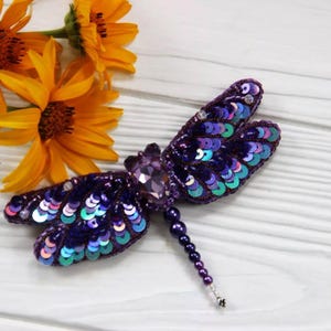 May include: A purple and iridescent dragonfly brooch made with sequins and beads. The dragonfly has a crystal-like body and a long beaded tail.