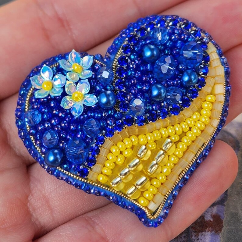 May include: A heart-shaped brooch made of blue and yellow beads, with a cluster of blue and white flowers on one side. The brooch is a symbol of support for Ukraine.