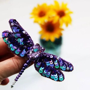 May include: A purple and blue dragonfly brooch made of iridescent sequins and beads. The dragonfly has a large, clear crystal in the center of its body.