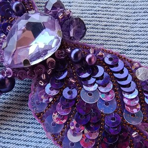 May include: A purple and iridescent brooch with a large crystal in the center, surrounded by smaller crystals and sequins. The brooch is made of fabric and has a textured surface.