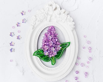 May include: A purple and green beaded lilac flower brooch in a white ornate frame. The brooch is surrounded by scattered purple and clear beads.