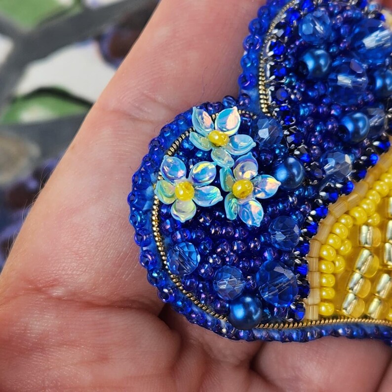 May include: A blue and yellow beaded heart-shaped brooch with a cluster of light blue flowers with yellow centers.