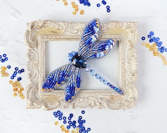 Pode incluir: Um broche de libélula azul e dourado feito de contas e lantejoulas, exibido em uma moldura de estilo vintage.