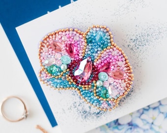 May include: A beaded brooch in shades of pink, blue, and green, with a gold-colored border. The brooch features various shapes of crystals and beads. A gold ring and necklace are also visible.
