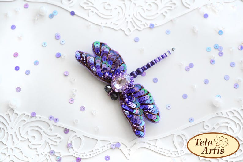 May include: A purple and blue dragonfly brooch made of sequins and beads. The dragonfly has a clear rhinestone in the center of its body. The brooch is on a white background with scattered sequins.