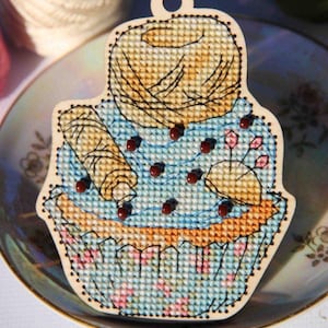 Może przedstawiać: Ozdoba w kształcie babeczki wykonana haftem krzyżykowym w kolorze niebieskim i żółtym, z kulką włóczki na górze. Babeczka jest ozdobiona brązowymi koralikami i różowymi kwiatami.