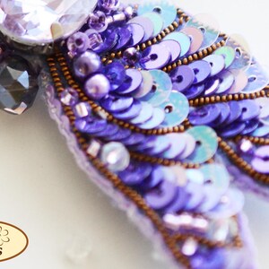 May include: A purple and blue sequined brooch with a large clear crystal in the center. The brooch is decorated with small purple beads and has a delicate, feminine design.