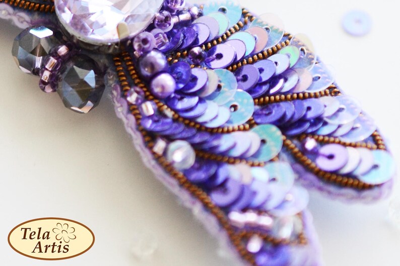May include: A purple and blue sequined brooch with a large clear crystal in the center. The brooch is decorated with small purple beads and has a delicate, feminine design.