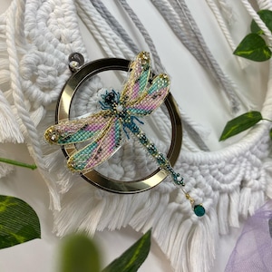 Op de afbeelding: Een gedetailleerde broche in de vorm van een libel met iriserende vleugels in tinten roze, groen en goud. De libel is bevestigd aan een gouden ring en heeft een hangende groene kraal.