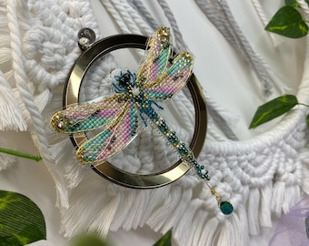 May include: A beaded dragonfly brooch with iridescent wings in shades of pink, green, and gold. The dragonfly is attached to a gold-colored ring and has a dangling green bead.