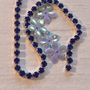 May include: A strand of blue rhinestone chain with a silver backing. The chain is in a circle shape with a straight piece extending from the circle. The chain is on a white surface.