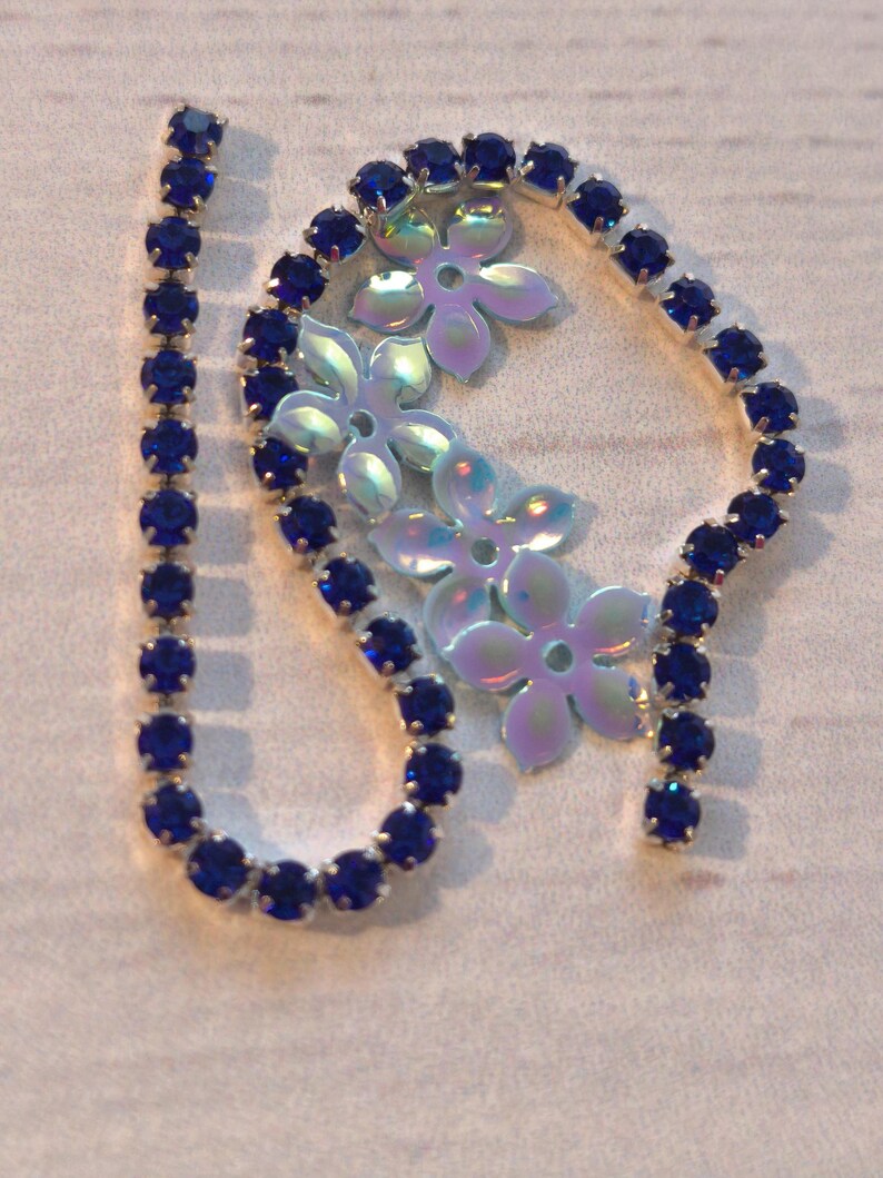 May include: A strand of blue rhinestone chain with a silver backing. The chain is in a circle shape with a straight piece extending from the circle. The chain is on a white surface.