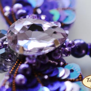 May include: A purple and blue dragonfly brooch with a large, clear crystal in the center. The brooch is made of beads and sequins.