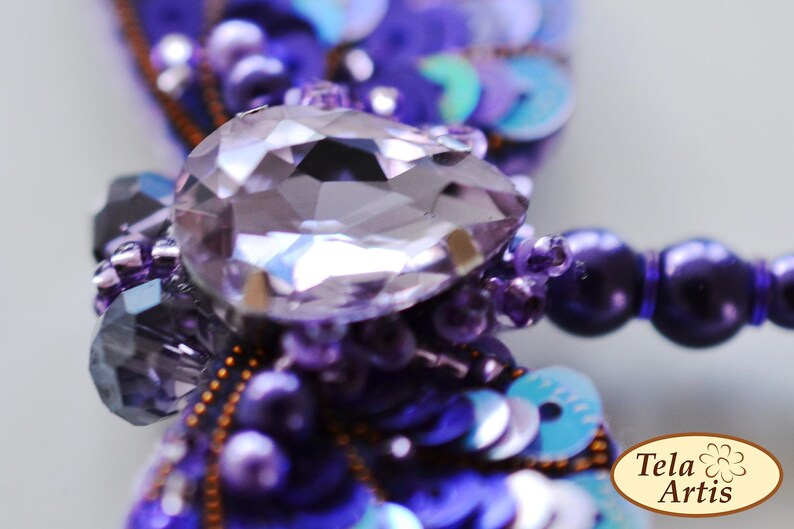 May include: A purple and blue dragonfly brooch with a large, clear crystal in the center. The brooch is made of beads and sequins.