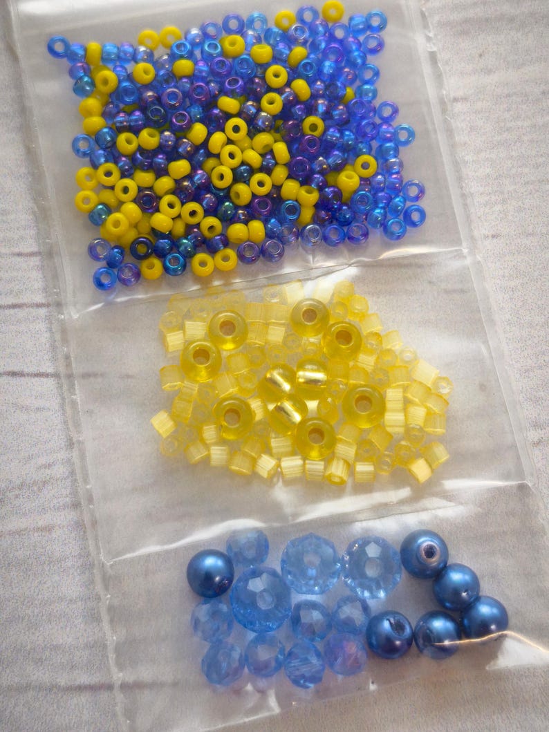 May include: Three bags of beads in blue and yellow colors. The first bag contains small round blue and yellow beads. The second bag contains small square yellow beads. The third bag contains larger round blue beads.