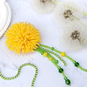Puede incluir: Una flor de diente de león de cuentas amarillas con tallos y hojas de cuentas verdes. La flor cuelga de una cadena de cuentas verdes. El fondo es una superficie blanca con dos flores de diente de león blancas.