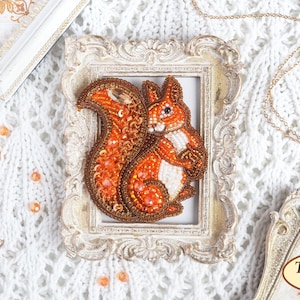 May include: A handcrafted squirrel brooch, meticulously beaded with orange, brown, and white beads. The squirrel is set in a decorative, ornate frame. The brooch is displayed on a white, textured surface with other decorative elements.