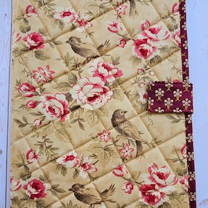 May include: A quilted fabric book cover featuring a beige background with a pattern of brown birds, green leaves, and pink flowers. The cover has a dark maroon border with a small floral print and a matching rectangular tab closure on the side.