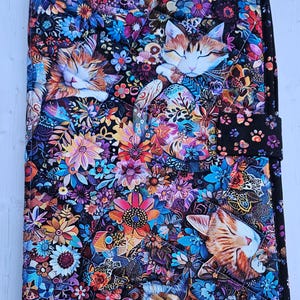 May include: A quilted notebook cover featuring a vibrant floral and cat design. The cover showcases a black background with colorful flowers in shades of blue, orange, purple, and pink. Sleeping cats are interspersed among the flowers. The cover has a black border and a snap closure.