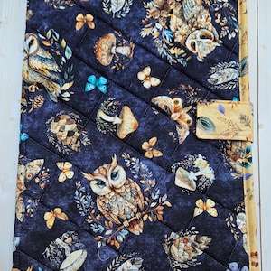 May include: A rectangular quilted fabric cover featuring a dark blue background with a repeating pattern of owls, mushrooms, butterflies, and feathers. The cover is finished with a mustard yellow border and a matching side tab closure.