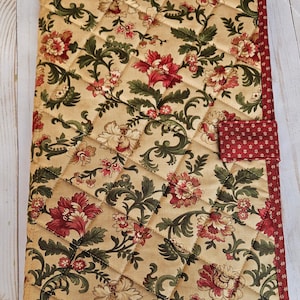 May include: Quilted journal cover with a floral pattern in red, green, and cream. The cover features a diamond-stitched design and a red border with a small dot pattern. A matching tab closure is on the right side.