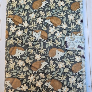 May include: A rectangular quilted fabric cover featuring a repeating pattern of brown hedgehogs, cream-colored flowers, and green leaves on a dark gray background. The cover has a light-colored border with a small tab closure on the right side.