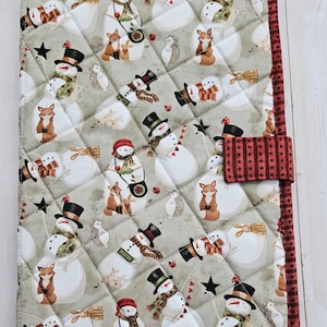 May include: A quilted fabric book cover featuring a light green background with a repeating pattern of snowmen, foxes, hedgehogs, and black stars. The cover is finished with a dark red border patterned with small black stars and a matching tab closure.