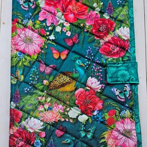 May include: A rectangular quilted fabric cover featuring a dark teal background with a vibrant print of pink and red flowers, green peacock feathers, and colorful butterflies. The cover includes a small side tab closure and diamond stitching.