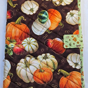May include: A rectangular quilted fabric cover featuring a dark brown background with a repeating pattern of orange, white, and green pumpkins and small leaves. The cover has a light green border with a leaf print and a matching side tab closure.