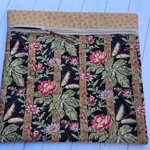 May include: Quilted fabric case with a floral pattern. The case features a black background with red, pink, and blue flowers, green leaves, and tan vertical stripes. A tan zipper and a brown leather pull are visible. The top border is tan with small floral designs.