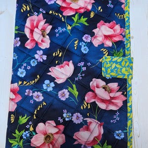 Op de afbeelding: Een rechthoekige, doorgestikte stoffen hoes met een donkerblauwe achtergrond en een bloemenpatroon van grote roze bloemen, kleine paarse en blauwe bloemetjes en groene bladeren. De hoes heeft een limoengroene rand met bladmotief en een sluitlipje.