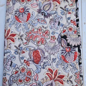 May include: Quilted floral pattern notebook cover in beige, red, blue, and purple. The cover features a repeating design of stylized flowers and leaves. The edges are trimmed with a black border and a black snap closure.