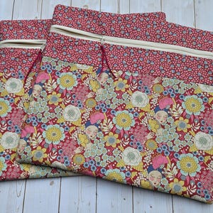 May include: Two rectangular fabric pouches featuring a zipper closure at the top. The pouches are made of a floral print fabric with stylized faces, yellow and white flowers, and pink accents, with a solid red patterned band at the top.