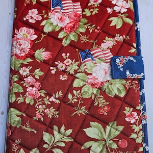 May include: Quilted notebook cover with a floral pattern in red, pink, and green, featuring American flags. The cover has a blue border and a blue tab closure with a floral design. The notebook cover is approximately 9 inches by 12 inches.