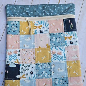 May include: A quilted zippered pouch with a patchwork design. The patchwork features squares of various patterns, including dragonflies, flowers, and ducks, in shades of blue, pink, and yellow. The top section is a solid blue fabric with a floral pattern.