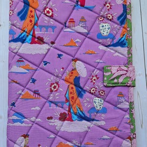 May include: A rectangular quilted fabric cover featuring a lavender background with a repeating pattern of figures in orange and blue robes holding parasols, small boats, pagodas, and floral motifs, finished with a green patterned border.