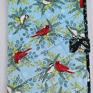 May include: A rectangular quilted fabric cover featuring a light blue background with a pattern of red cardinals, black-capped chickadees, green pine branches, and white snowflakes. The cover has a black border and a small black tab closure.