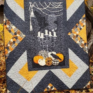 May include: A Halloween-themed quilt with a dark gray background and geometric patterns in silver and orange. The center features a black panel with a candelabra, pumpkins, and a crow. The quilt is displayed outdoors.