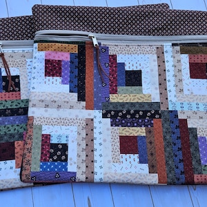 May include: Three rectangular quilted pouches with a log cabin design. The pouches feature a variety of fabrics in shades of brown, beige, purple, and red. Each pouch has a zipper closure and a brown fabric top.