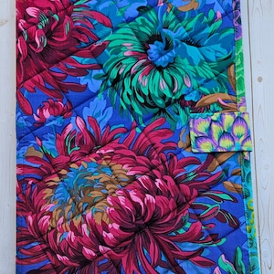 May include: A rectangular quilted fabric cover featuring a vibrant floral pattern with large magenta, teal, and green chrysanthemum-like flowers on a blue background, finished with a green border and a small matching fabric tab closure.
