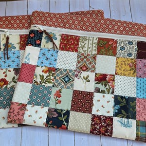 May include: Two patchwork zipper pouches with a variety of floral and geometric patterns in red, blue, and cream. The pouches have a red patterned top and a cream zipper with a leather pull. The patchwork design is made of small squares.