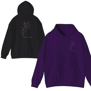 Puede incluir: Se muestran dos sudaderas con capucha, una negra y otra morada. Ambas tienen un bolsillo delantero y una capucha. La sudadera negra tiene un diseño de mariposa dorada en la espalda y el texto "butterfly giggle". La sudadera morada tiene el mismo diseño en la parte delantera.