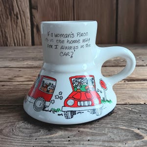 May include: White ceramic mug with a unique shape, featuring a handle and a wide base. The mug has a cartoon illustration of two red cars and the text: "If a woman's Place is in the home WHY AM I Always in the CAR?"