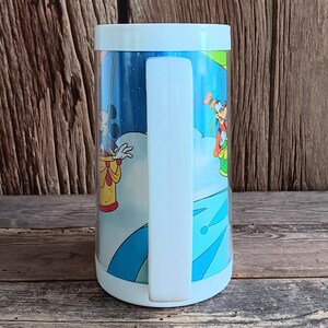 May include: A vintage, cylindrical mug with a white handle and rim. The mug features a colourful cartoon scene with Mickey Mouse and Goofy in a hot air balloon against a blue sky background. The mug is made of a clear, durable material.