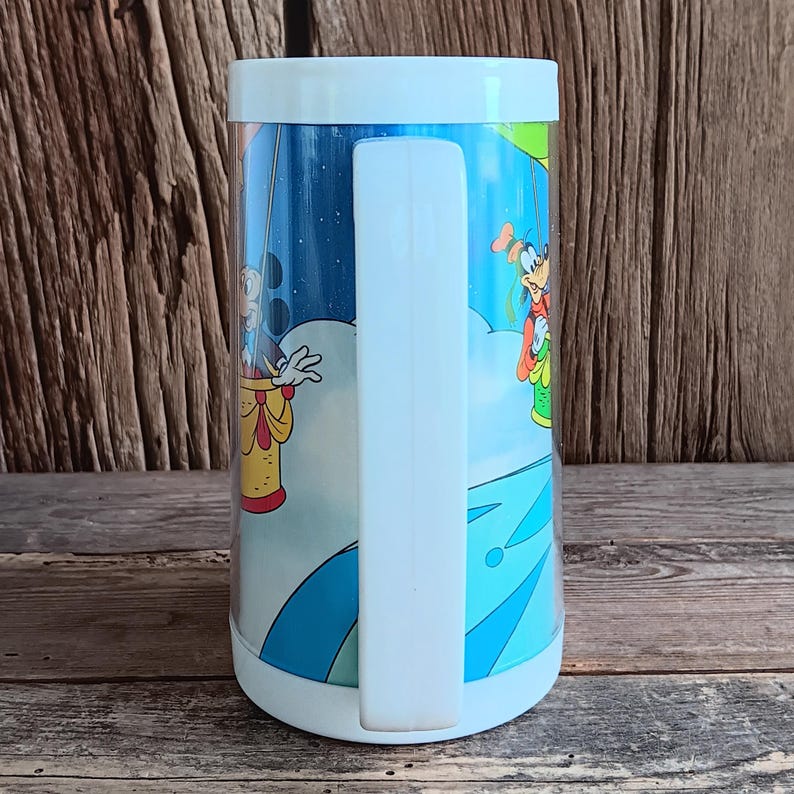 May include: A vintage, cylindrical mug with a white handle and rim. The mug features a colourful cartoon scene with Mickey Mouse and Goofy in a hot air balloon against a blue sky background. The mug is made of a clear, durable material.