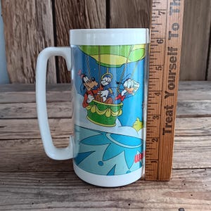 May include: A rectangular white mug with a colourful image of Goofy and Donald Duck in a hot air balloon against a blue sky. The mug has a white handle and rim. The mug is approximately 15 cm tall.