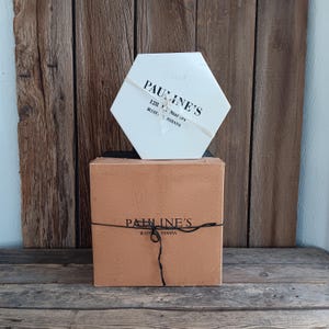 May include: A white hexagonal box with black text that reads "PAULINE'S 128 W. Market Bluffton, Indiana" is stacked on top of a brown box with black text that reads "PAULINE'S Bluffton, Indiana".